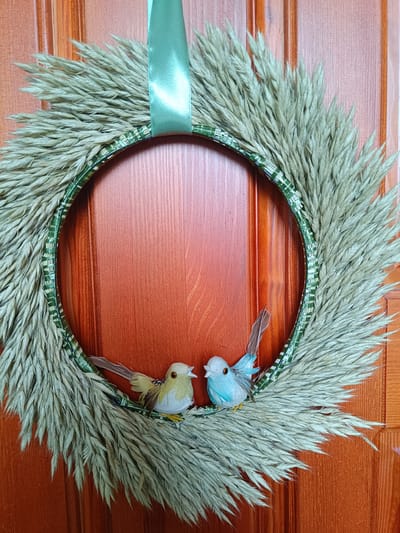 A wreath made of dried grass adorned with two small decorative birds hangs on a wooden door in Pāvilosta, Latvia. The wreath is circular and appears to be constructed around a green base. A green ribbon is used to suspend the wreath. One bird is light yellow and white, with brown accents, and the other is light blue with white accents. Both birds are perched on the lower portion of the wreath. The door is made of wood with a reddish-brown stain and vertical paneling.