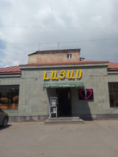 The image captures a storefront in Nor Hachin, Armenia, under a cloudy sky. The building features a light green, textured facade with a darker, checkered base. Above the entrance, a yellow neon sign displays the Armenian letters "ԼԻԶԱ" (Liza). To the right of the sign is a black rectangular plaque with a pink heart outline containing some text. A dark awning stretches above the entrance. To the left of the entrance, a large window showcases what appears to be decorative items or merchandise. A parked car is visible on the left side of the image, and to the right of the entrance, a small kiosk or machine stands near the doorway. The overall scene suggests a commercial establishment on a street in the city.