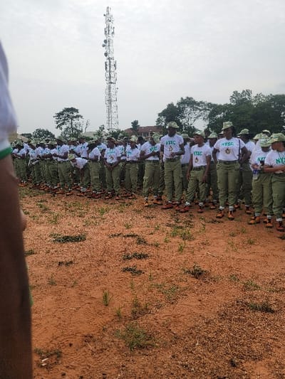 A long line of people in white shirts, green pants, hats, and brown boots stand in formation. The shirts have "NYSC" printed on them. The people are standing on red dirt. A tall communications tower stands behind them. The sky is overcast and gray. Some trees are also visible in the background.