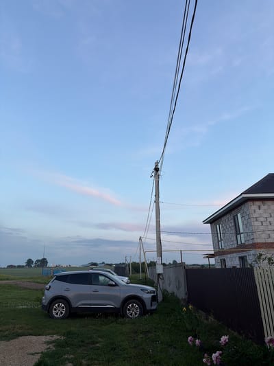 A gray SUV is parked on a grassy area next to a dark metal fence. A house under construction, made of concrete blocks, is visible behind the fence. A utility pole with wires runs diagonally across the frame. The sky is a gradient of blue and pink, suggesting it is either sunrise or sunset. The background shows a rural landscape with some distant buildings and trees. The scene appears to be in the city of Nizhniy Suyk-Su, Russia.