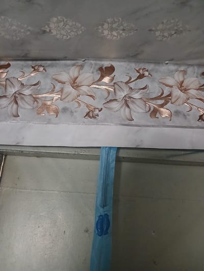The image shows a close-up of a ceiling and a portion of a wall. The ceiling has a decorative pattern of floral motifs in a light color against a marbled background. Below the ceiling is a floral border with lilies and other flowers in shades of beige and gold. This border is on a marbled background as well. Below the floral border, there is a white horizontal band, possibly trim or a different material. Further down, a blue zipper is visible, hanging vertically. The background below the zipper appears to be a wall with a textured surface, possibly painted or covered in a fabric. The lighting suggests an indoor environment. The location is specified as Hlushky, Ukraine.