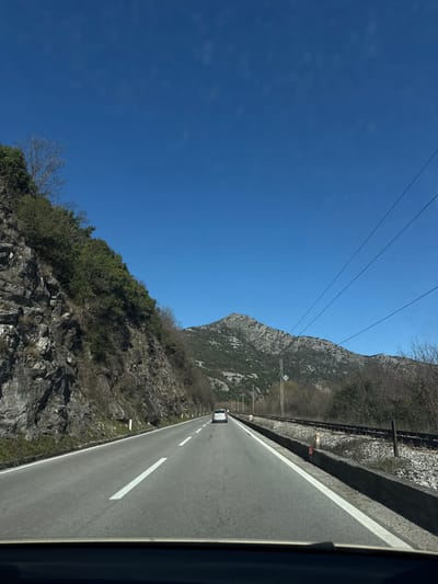 The image captures a forward-facing view from inside a vehicle on an asphalt road in Braćeni, Montenegro. A clear, deep blue sky occupies the upper portion of the frame. To the left, a steep, rocky cliff face rises, covered with a mix of evergreen and deciduous vegetation. White traffic delineators with red tops are visible along the left road shoulder. The road features a solid white line on the right shoulder and a dashed white line indicating the center. A light-colored sedan (silver or white) is visible ahead, traveling in the same direction. To the right of the road, separated by a low concrete barrier, a single set of railroad tracks runs parallel. Sparse, mostly barren vegetation lines the area beside the tracks, and a utility pole supports three overhead power lines that traverse the upper right quadrant. In the background, prominent, rugged mountains with visible rock formations and sparse vegetation extend across the horizon. The dark dashboard of the originating vehicle is visible at the bottom of the image. No individuals are depicted.
