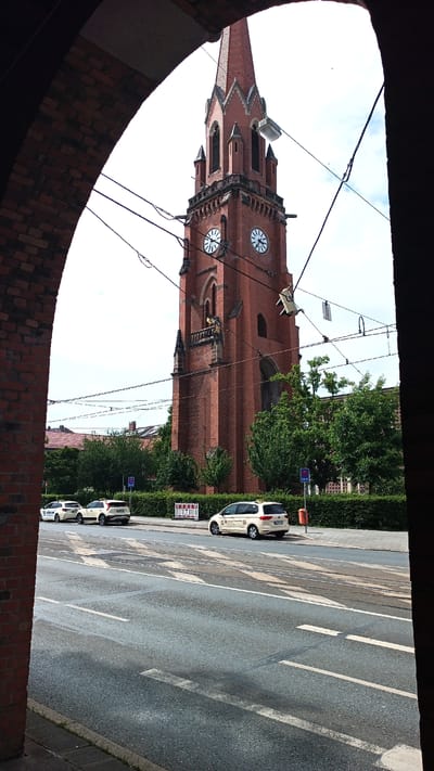 A brick archway frames a view of a red brick church tower with a tall spire. The tower has two clock faces, both showing approximately 3:00. Below the clock faces, there is a balcony with ornate detailing. Wires and a streetlight are visible in front of the tower.
In the foreground, a paved street runs horizontally across the image. Several white cars, some of which appear to be taxis, are parked along the side of the street. A green hedge lines the sidewalk behind the cars. In the background, buildings and trees are visible. The sky is overcast.
The scene appears to be set during the daytime, with the lighting suggesting it could be late morning or early afternoon. The presence of the cars and the general street scene suggest an urban environment. No people are clearly visible.
There is text on the side of one of the white cars, which appears to be a taxi. The text is in German. Another white vehicle has the word "TAXI" visible on its roof. A blue sign with "TAXI" written on it is also visible near the curb.