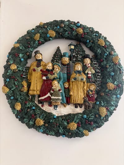 This media file features a highly detailed, three-dimensional Christmas wreath displayed against a plain white background, likely a wall. The circular wreath is crafted from sculpted material, possibly resin, and painted in rich, festive colors. The main body of the wreath is dark green, mimicking evergreen foliage, and is abundantly decorated with golden-brown pinecones, clusters of small red berries, and interspersed light blue/teal spherical ornaments.
Within the wreath's open center, a charming winter scene unfolds. A group of stylized figures, appearing to be people from a bygone era (possibly Victorian carolers), are arranged vertically. They wear warm, layered winter clothing, including long coats in shades of gold, red, blue, and olive green, complemented by hats, bonnets, and scarves. Several figures are depicted holding small, open books, suggesting they are singing carols. One smaller figure holds what appears to be a doll. The background of this inner scene includes miniature evergreen trees lightly dusted with white, sculpted snow, which also covers the ground around the figures.
The figures have simple, serene expressions, contributing to a nostalgic and festive atmosphere. The overall piece evokes a traditional Christmas or winter holiday gathering. There is no visible text in the image. The location context for this media file is Chatham, United Kingdom.