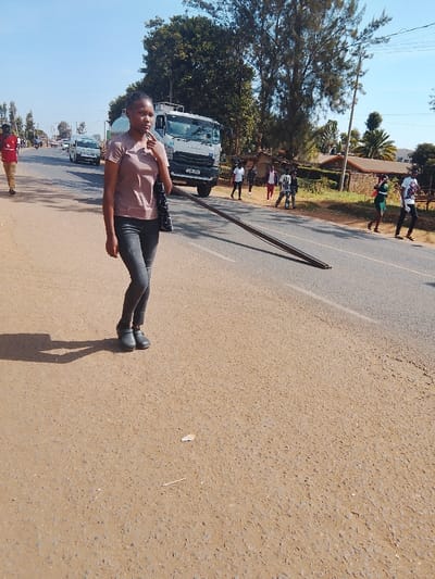 A young woman in a short-sleeved mauve shirt and dark jeans walks along a paved road, carrying a black purse. Behind her, a white tanker truck is parked near the side of the road, and several people are walking in the distance on the other side of the road. There is a long, dark pole that stretches across the width of the road. Trees and buildings are visible in the background. The sky is clear and blue.