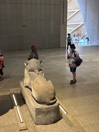 In the dimly lit, modern interior of a museum hall, a stone statue of a seated lion rests on a pedestal. Two individuals are present. In the foreground, slightly to the right, a person wearing a red fez, a patterned tunic, and denim shorts stands looking at a camera, holding it in their hands. To the left and further back, another person also wears a red fez and a traditional vest over a lighter garment. In the background, a man in a light blue jacket is seen looking at his phone, and beyond him, a crowd of people is gathered in a large, open space with angular architecture and high ceilings. The floor is tiled with a speckled pattern, and vertical light fixtures are embedded in the floor near the statue. The overall environment suggests an indoor exhibition space with contemporary design elements. The location is the Grand Egyptian Museum, located near Kafrat Nassar, Egypt.