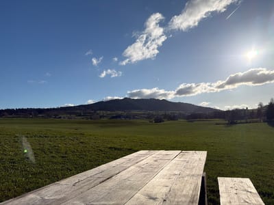 The image captures an expansive outdoor landscape in Gibloux, Switzerland, under clear, sunny conditions. In the immediate foreground, a weathered, light-colored wooden picnic table occupies the lower portion of the frame, extending from the bottom edge and comprising distinct horizontal planks. A disconnected section of a matching wooden bench is visible to the right of the table. Beyond the table, a vast, undulating green grassy field stretches across the midground. The background is dominated by a prominent, densely forested hill or mountain range, exhibiting dark green foliage. At the base of this elevation, distant, faint structures suggest a small settlement. The sky above is a bright, unclouded blue, with several scattered white cumulus clouds concentrated in the upper right quadrant. A bright sun is visible in the upper right corner, producing a notable lens flare, and a white contrail from an aircraft traverses the upper right sky. No individuals, actions, or specific interactions are present in the scene.