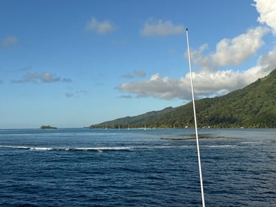 The image presents a panoramic coastal scene in Fa'a'ā, French Polynesia, under a bright blue sky dotted with scattered white cumulus clouds. The perspective is from a low vantage point, indicated by a white pole with a black tip extending diagonally from the bottom center-right towards the upper center-right of the frame. The foreground features choppy, dark blue ocean water with a prominent white-capped wave breaking horizontally across the mid-left portion. In the midground, a small, densely vegetated islet is visible on the far left, surrounded by lighter blue, calmer waters. Along the distant shoreline to the right, several white sailboats are anchored or moored in a lagoon characterized by varying shades of light blue and turquoise, indicative of shallow areas or reef structures. The background is dominated by a long, steep, and heavily forested mountain range covered in lush green tropical vegetation. Scattered small, white architectural structures are discernible at the base of the mountains along the coast. No individuals are visible.