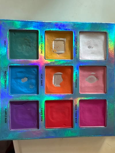 The image displays a nine-pan eyeshadow palette featuring a holographic, iridescent cardboard exterior. The pans are arranged in a 3x3 grid. In the top row, from left to right: the first pan contains a teal shade labeled 'JADED' with minor surface use; the second pan, labeled 'HELLO SUNSHINE', contains a yellow shade heavily depleted in the center, exposing the silver pan; the third pan, labeled 'COCONUT CRUSH', contains an off-white shade with light surface marks. In the middle row, from left to right: the first pan, labeled 'PACIFIC DREAMS', contains a bright blue shade with a small circular area of product missing, revealing the pan; the second pan, labeled 'HEAT WAVE', contains a bright orange shade with a large, irregularly shaped portion removed, exposing the pan; the third pan, labeled 'PINKY PROMISE', contains a light pink shade with an elongated oval depression, revealing the pan. In the bottom row, from left to right: the first pan, labeled 'PURPLE RAIN', contains a deep purple shade with minimal signs of use; the second pan, labeled 'CARPE DIEM', contains a red shade with minimal signs of use; the third pan, labeled 'MAKE THEM WINK', contains a hot pink shade with minimal signs of use. The palette is situated on a plain, light-colored surface. There are no discernible features in the image to indicate its location in the city of Plasencia, Spain.