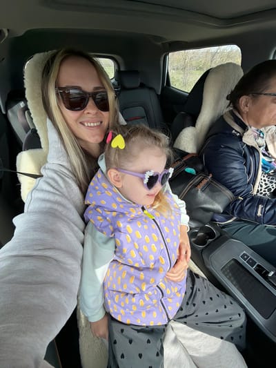 The image shows three people inside a car. A woman in sunglasses is in the foreground smiling, with her head resting on a sheepskin car seat cover. A young girl wearing a purple and yellow patterned jacket and sunglasses is lying down next to her. Behind them, a third person is seated, wearing glasses and a blue jacket. The car's interior is visible, including the center console and a window showing an outdoor scene with trees. The location, Ternovka, Russia, is not visually confirmed in the image.