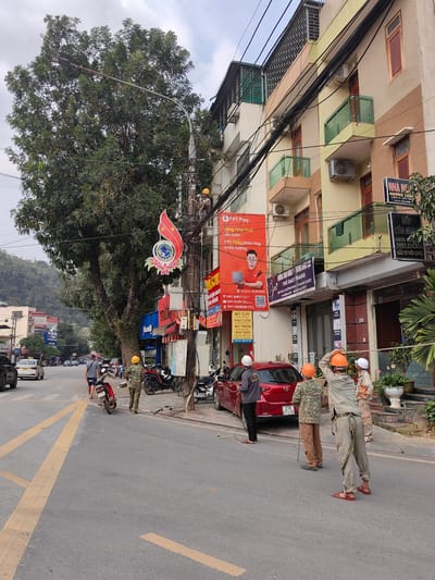 The image depicts a street scene in Thành phố Hà Giang, Vietnam. Several utility workers wearing helmets are gathered on the side of the road, observing and working on an electrical pole. One worker appears to be on the pole. There is a red car parked nearby, with license plate 23A 074.56. Other cars and motorcycles are also visible on the street, as well as pedestrians walking. Several buildings with storefronts line the street, with Vietnamese signage on them. The foliage of a large tree is seen in the upper left of the image. The sky is overcast. The scene appears to be an urban setting with a mix of residential and commercial buildings.