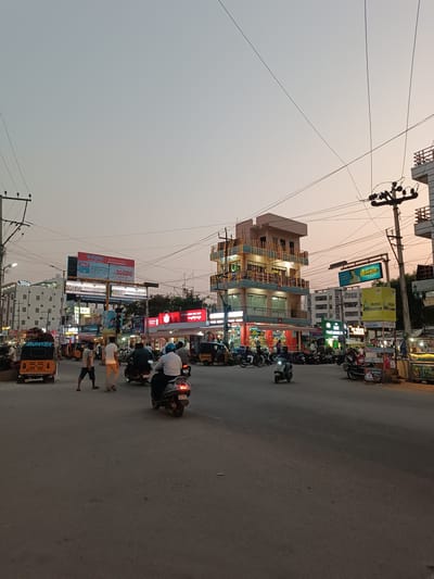 This image depicts a bustling urban street scene in Anantapur, India, at dusk or early evening, characterized by artificial illumination against a fading sky.

The sky exhibits a gradient from light grey overhead to a pale orange-pink near the horizon. Numerous dark overhead electrical and communication wires crisscross the upper portion of the frame, connected to multiple utility poles.

The foreground features a dark asphalt road, generally clear but showing some surface irregularities.

Along both sides of the street, multi-story buildings house various commercial establishments and possibly residences. On the far left, a white building labeled "KOMALA LODGE" is visible. In the center-right, a prominent three-story building, light blue and white, is adorned with numerous strings of yellow-white decorative lights. Its ground floor features illuminated storefronts, including one with a red and white striped awning. Further right, additional buildings and vendor stalls with illuminated displays line the street.

Multiple large billboards and smaller signs are visible throughout the scene. Notable billboards include "Reliance Digital," "Anantapur's Best Big School," "Manju Granites," "Ramana's," and "Muttabally Mall," confirming the city as Anantapur.

Vehicular traffic is moderate. Numerous two-wheelers (scooters and motorcycles) are present; some are in motion, and many are parked along the curbs. In the immediate foreground, a person wearing a light shirt and blue helmet is riding a dark scooter away from the camera, with its taillight illuminated. Another lighter-colored scooter is moving left in the mid-ground. At least two yellow auto-rickshaws are visible, one prominently on the far left, labeled "HUNTER," facing right.

Approximately 10-15 individuals are present. Two male pedestrians are walking on the road in the center-left, one in a light t-shirt and dark shorts, the other in a white t-shirt and khaki pants. The remaining individuals are mostly operators or passengers of two-wheelers. Actions primarily involve commuting and street-level commerce, indicated by active shops and vendor stalls.