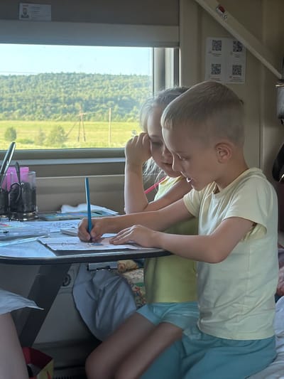 Two children, a boy and a girl, are drawing at a table on a train. The boy, on the right, is wearing a yellow t-shirt and blue shorts and is actively coloring. The girl, on the left, is wearing a yellow shirt and is leaning her head on her hand, watching the boy. The table is cluttered with drawing supplies, including papers, a blue crayon, and what appears to be a watercolor set. Through the window behind them, a green, hilly landscape with trees is visible, suggesting the train is traveling through a rural area. The interior of the train car is dimly lit, with beige paneling and some decorative stickers on the wall. The scene suggests a quiet, indoor activity during a train journey, possibly in or near the city of Mozhga, Russia, given the context.