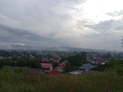 The image shows a landscape view of Uruekanoe, Indonesia, under an overcast sky. In the foreground, tall grass obscures the lower portion of the scene. Beyond the grass lies a cluster of buildings, including residential houses with a mix of red and gray rooftops. The buildings are interspersed with trees. In the background, a mountain range is visible, shrouded in a light mist. The sky is predominantly gray with some lighter areas suggesting a break in the cloud cover. No people are visible in the frame.