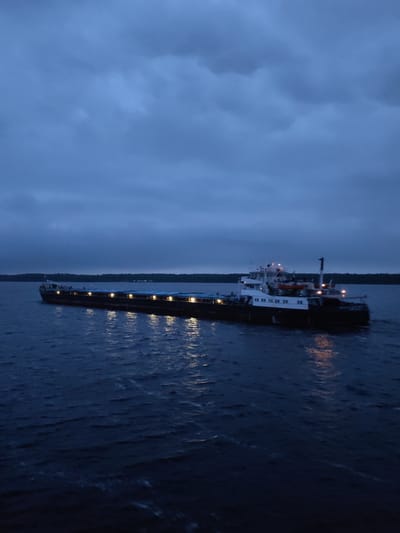 A long, dark barge with a multi-tiered white superstructure is traveling on a body of water under an overcast sky. The barge has numerous lights illuminated along its length, reflecting in the water. Dark vegetation appears on the shoreline in the distance. The photo was taken in Voznesenye, Russia.