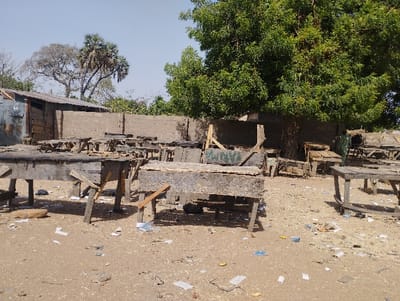 The image shows a scene of a market area in Kongo, Nigeria. The scene is littered with debris and discarded items, including small pieces of paper and plastic. The ground is dry and dusty. The image is taken on a sunny day. There are wooden benches in the center of the image. Some are damaged and broken. There are trees and buildings in the background.