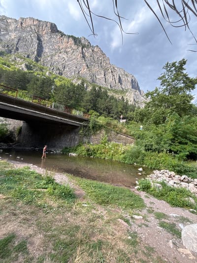 In a natural setting with a mountain in the background and lush greenery surrounding a shallow stream, a person is wading in the water. The stream flows from left to right in the frame, with a bridge crossing over it. The bridge has a concrete structure and a red and white metal railing. On the bank of the stream, there are rocks and grass, with a dirt path alongside. The mountain face is rugged and rocky, with trees growing on its slopes. The sky is visible, with some clouds. The person in the stream is wearing swimwear and appears to be enjoying the cool water. There are no visible signs or text. The scene suggests a warm day, possibly in the summer, given the presence of the person in swimwear and the green foliage. The location is identified as Zgorigrad, Bulgaria.