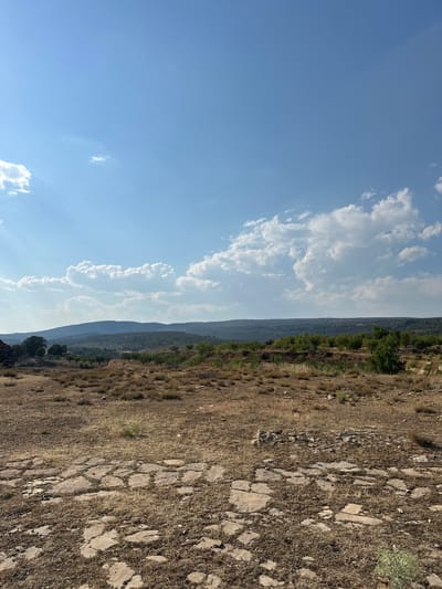 The image shows a dry, arid landscape with rolling hills and a clear blue sky dotted with white clouds. In the foreground, there is a patch of rough, dry ground with scattered rocks and dried vegetation. A portion of the ground appears to be paved with irregular, flat stones, suggesting a historical or ancient pathway or structure. The middle ground features more dry, sparse vegetation and a few scattered trees. In the background, a series of undulating hills, covered in green and brown vegetation, stretch across the horizon. The overall scene depicts a natural, somewhat barren environment under a bright sky. The location is identified as Alpuente, Spain. There are no people or animals visible in the image.