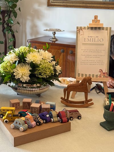 This photograph, taken in La Molina, Peru, captures a table setting. The focal point is a floral arrangement featuring white dahlias and green hydrangeas in a decorative silver bowl. Beside the arrangement, there is a wooden rocking horse. Adjacent is a vertical decorative display of wooden blocks spelling "BABY" along with stylized cars. To the left, a framed sign on a small easel reads "BABY CAR EMILIO". The text on the sign is in Spanish and provides instructions for decorating a toy car for Emilio. On the table there is a container with writing utensils and a partially visible plate with pastries. In the background is a wooden cabinet. The lighting suggests an indoor setting, possibly for a baby shower or related event.