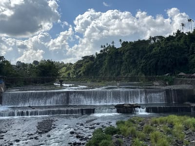 This image shows a tiered waterfall and dam structure on a river, with lush green hills and palm trees in the background under a cloudy blue sky. In the distance, a bridge spans the river. Several small structures, possibly huts, are visible on the riverbank near the top of the dam. The riverbed is covered with stones and rocks, and the water flows over the man-made tiers, creating a cascading effect. The environment suggests a tropical or subtropical location. Based on visual cues, this appears to be the Tukad Unda river dam in Klungkung, Bali, Indonesia, a known location for its multi-tiered waterfalls. There are no people clearly visible in the image.