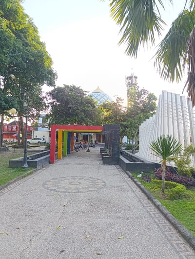 A paved walkway with a stone pattern leads to a colorful metal archway, located in Kecamatan Asembagus, Indonesia, at latitude: -7.749, longitude: 114.218. The archway is surrounded by green trees and shrubs. A white mosque with a dome is visible in the background, alongside a white tower structure. On the right side of the scene, a white wall structure rises up with a small palm tree in front of it. There are several parked motorcycles behind the archway. The scene appears to be a public park with an outdoor space for gathering. The weather is sunny with clear blue skies.