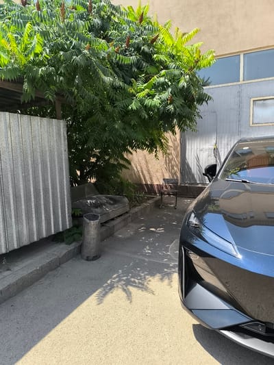 The image depicts an outdoor scene in a paved area, possibly a parking lot or alleyway, with a dark blue car in the foreground. To the left of the car, there is a tall structure made of corrugated metal sheeting, adjacent to a patch of greenery including a large tree with dense foliage. A discarded, dark-colored sofa or seating structure rests on a concrete border near the metal sheeting, alongside a cylindrical metal object that could be a bin. Further into the background, a building with a light-colored facade is visible, featuring a large window and a grey metal door. A simple chair is placed near the wall. The scene is illuminated by sunlight, casting shadows from the tree onto the ground and car. The location is identified as Іргелі ауылдық округі, Kazakhstan. No people are present in the image.