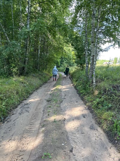 In the foreground of the image, a sandy dirt road curves through a wooded area. The road exhibits tire tracks, indicating it is a path used by vehicles. On the left side of the road, tall grasses and bushes grow, with a dense forest of trees beyond. On the right side, the vegetation is also present, with birch trees prominently featured.
Two individuals are walking away from the camera down the dirt road. The person in front is wearing a light blue hooded jacket, gray pants, and red boots. They appear to be carrying something in their right hand. Behind them, a second person is wearing a dark green jacket and dark pants, and they are carrying a green bag in their left hand.
The scene suggests a walk or hike in a rural or semi-rural environment. The presence of trees and dense vegetation implies a natural setting. The location is specified as Verkhneye Kazachye, Russia.
