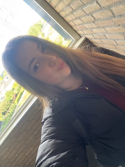 The media file is a selfie of a young person with long, light brown hair. They are wearing a dark jacket over a dark red top and a small, delicate necklace. The subject's face is turned towards the camera, and they appear to be looking slightly off-camera. Their expression is neutral.
The setting appears to be indoors, near a window. Through the window, greenery and trees are visible, suggesting an outdoor environment. The sun is shining brightly, casting a strong light on the scene, particularly on the window and the subject's face, which creates some lens flare. A brick wall is visible to the left and right of the frame, indicating the building's construction.
The photo is taken in Enschede, Netherlands. The overall impression is of a casual, everyday moment captured by the subject themselves. The time of day is likely daytime, given the bright sunlight. There is no visible text in the image.