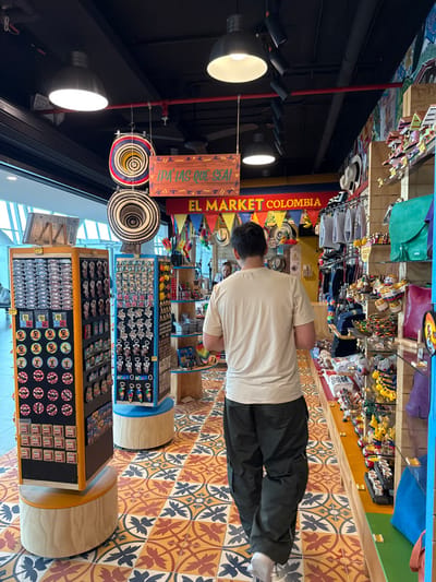 An interior view depicts a souvenir shop, identified by a banner reading "EL MARKET COLOMBIA" and another sign stating "¡PA' LAS QUE SEA!", located within Colombia, specifically inferred to be in the Area Metropolitana Centro Occidente (AMCO). The shop features a black exposed ceiling with red piping and multiple industrial-style pendant lights and track lighting fixtures. The floor is tiled with a distinctive pattern of orange, yellow, red, and black geometric and floral motifs.
A male individual, viewed from behind, occupies the foreground and middle ground, walking away from the camera towards the rear of the store. He wears a light beige short-sleeved t-shirt, dark olive-green cargo pants, and light-colored footwear. His dark hair is short. A blurred figure is partially visible deeper within the shop, to the left of the walking individual.
The shop is extensively stocked with merchandise. On the left, two tall, black and yellow rotating display stands present numerous keychains, magnets, and decorative pins. Hanging from the ceiling are two traditional Colombian "sombrero vueltiao" hats and one white fedora-style hat. To the right, a long, multi-tiered shelving unit extends into the background, filled with various souvenir items including small figures, textile bags, and other crafts. Colorful triangular flags (red, yellow, blue) are strung above the main "EL MARKET COLOMBIA" banner. The left side of the scene reveals a large window or glass partition, indicating an open-plan environment.