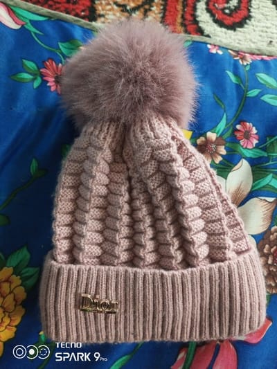A dusty rose knitted winter hat is positioned on a vibrant blue fabric background adorned with a floral pattern, featuring large white, pink, and yellow flowers with green leaves. The hat itself has a chunky, textured knit body, a ribbed brim, and is topped with a large, fluffy pom-pom of a matching color. A small, metallic gold-colored "Dior" logo is affixed to the front left portion of the hat's brim. Portions of a red and cream patterned textile are visible in the upper background. A "TECNO SPARK 9 Pro" watermark is present in the lower left corner of the image. The scene is located in the city of Khidirsho, Uzbekistan.