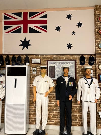 The image displays a room showcasing Australian naval history. A large Australian flag hangs on the left wall, decorated with stars. Three mannequins dressed in navy uniforms stand on the right, representing different ranks. Behind the middle figure, a framed "Campaign & Battle Honours" poster is visible. An air conditioner is placed in the bottom right corner, and naval caps are displayed along the wall. Additional naval decorations are present on the upper wall, and historical documents are framed on the wall above the air conditioner.