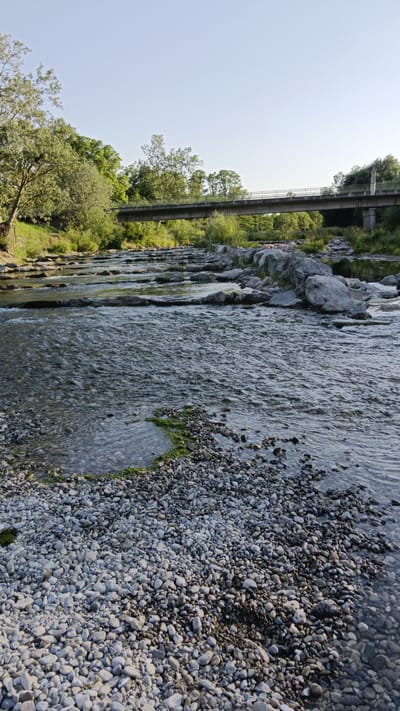 This is a vertical, eye-level, medium shot of a shallow river flowing over rocks and gravel. In the foreground, a stony riverbank is covered in small, light-colored pebbles. The river water is clear, allowing the submerged rocks and rippling surface to be visible. Several flat stones are laid across the river, creating small waterfalls. In the background, a concrete bridge with a metal railing spans the river, surrounded by lush green trees and vegetation. The sky is clear and pale blue, suggesting it is daytime, likely late afternoon or early evening due to the soft light and warm tones. The overall mood is peaceful and serene, evoking a sense of nature and tranquility. No people or text are visible in the image. The scene is consistent with a natural setting in Sankt Pölten, Austria, known for its rivers and green spaces.