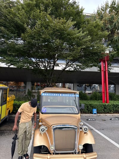 The image captures an outdoor scene in the Olympic Sailing Marine Culture Tourist Area, Shinan District, Qingdao, China. A male individual, appearing to be of Black ethnicity, stands in the foreground, positioned to the left of a gold-colored, vintage-style electric tourist vehicle. He wears a beige short-sleeved polo shirt, olive-green cargo pants with white drawstrings, black slide sandals, and a black flat-brimmed cap. He holds a folded black umbrella loosely in his left hand, and his head is tilted downwards, directed towards the front of the gold vehicle.
The gold vehicle is a four-wheeled, open-top electric tourist transport, featuring a chrome grille, circular headlights, and a windshield banner inscribed with "奥帆海洋文化旅游区" (Olympic Sailing Marine Culture Tourist Area). A blue circular sign displaying the number "7" is affixed below the windshield on the driver's side. Partially visible on the far left, behind the man, is a portion of a yellow utility or golf-cart style vehicle with a black soft top.
The background features a large deciduous tree with dense green foliage, obscuring part of a modern, multi-story building characterized by dark grey paneling and reflective windows. To the right of the gold vehicle, a patch of green shrubs precedes a tall, slender red architectural structure. The ground consists of asphalt pavement marked with white lines, and black curb stoppers are visible on the right. A small blue sign with a white ship emblem is positioned near the curb stoppers.