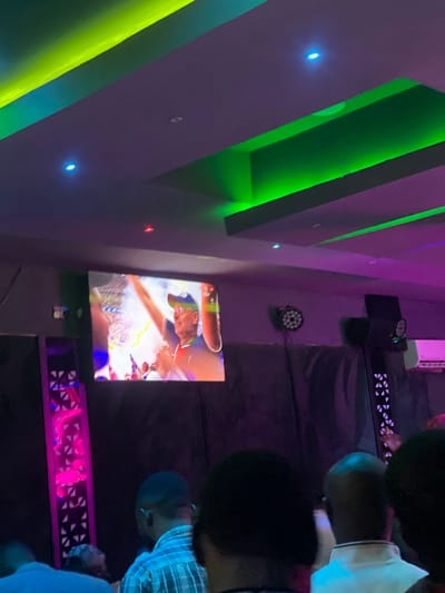 The image shows a group of people watching a television screen in a room with purple walls and green and yellow lighting fixtures. The TV screen is displaying a person in a golf uniform with their arms raised in the air. The setting is likely a bar or lounge, based on the dim lighting and the presence of people gathered together. There is no information to verify the image's location in Rubuchi, Nigeria.