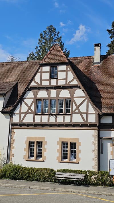The image shows a building in Rheinfelden, Switzerland. The building has a red-tiled roof and a white facade with dark brown timber framing on one part. The rest of the building has square windows with brown trim. There is a bench located alongside a hedge, and a street runs next to the hedge. The sky is blue with some clouds. Part of a tree is visible on the left side of the building. The picture is taken on a sunny day.