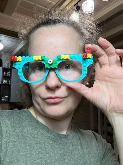 A person is holding up a pair of glasses made of colorful LEGO bricks in front of their face. The glasses have a blue frame with red, yellow, and green LEGO pieces attached to the sides. The person is wearing a green shirt and has short curly hair. There are building materials in the background. The image was likely taken in an indoor environment. It is not possible to determine the exact location in Центроспецстроевец Снт, Russia, from the given image.