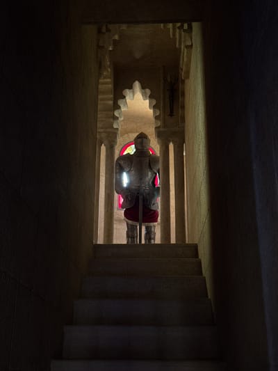 The image depicts a dimly lit, enclosed space with stone architecture. The primary subject is a statue of a knight in full armor, lying horizontally with a red cloak. The statue is illuminated by a bright light that reflects off its metallic surface. Behind the statue, a stained-glass window with red and yellow panes is visible. The setting appears to be a crypt or memorial, characterized by the rough-hewn stone walls and tiered structures that suggest stairs or seating. The overall atmosphere is somber and reverent, with the shadows and focused lighting creating a dramatic effect. The image was taken in Benalmádena, Spain, which could indicate a historical or religious significance to the site. There is no visible text in the image.