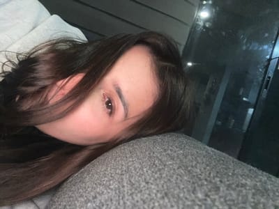 A woman with long, dark hair lies down, her head resting on a gray, textured surface. Her face is partially visible, featuring one eye and part of her forehead. The background reveals a window reflecting city lights or street lamps, suggesting an urban environment. The overall lighting is dim, creating a soft and slightly melancholic atmosphere.