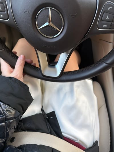 The image shows the interior of a car, specifically the steering wheel of a Mercedes-Benz. The dashboard and a seat are visible behind the steering wheel. A hand is gripping the steering wheel. The seat belt is buckled. A jacket is partially visible on the right. The exact location of the photo is not discernable in the image.