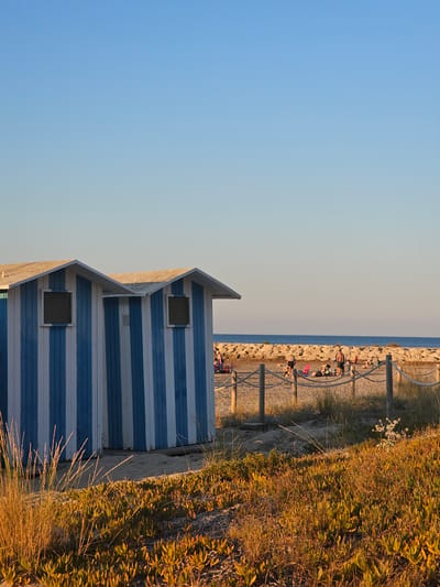 This image depicts a beach scene in Deveses, Spain, bathed in the warm glow of late afternoon or early morning sunlight. In the foreground, two beach huts with blue and white vertical stripes stand side-by-side. Each hut has a simple, pitched roof and a small, square window. Adjacent to the huts, sandy ground is covered with low-lying green and yellow vegetation, with tall, dry grass in the immediate foreground. A rope fence with wooden posts separates the huts from the beach area beyond.
In the midground, several people are scattered on the sand, some reclining on chairs and towels, others standing or walking. They appear to be engaged in typical beach activities like sunbathing or relaxing. A rough, stone breakwater runs horizontally across the scene, separating the beach from the calm, blue sea. The water stretches to the horizon under a clear, pale blue sky. The overall atmosphere is tranquil and summery.