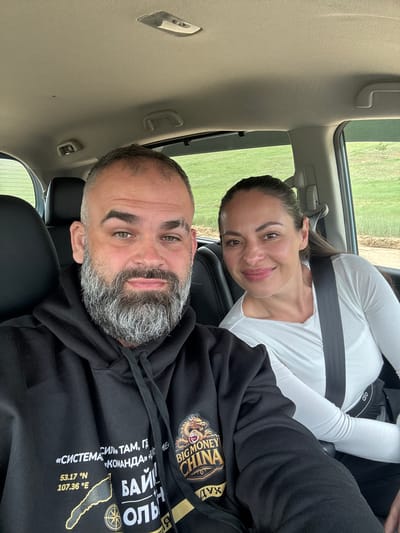 The image is a close-up selfie taken inside a car. A man with a gray beard and dark hoodie is in the foreground, with his face turned towards the camera and a slight smile. Next to him, a woman with dark hair and a white long-sleeved shirt is also looking at the camera and smiling. Both are wearing seatbelts. The background shows a green, hilly landscape, consistent with the outskirts of a town. The text on the man's hoodie is partially visible, featuring Cyrillic script and what appears to be a dragon emblem with the words "BIG MONEY CHINA". The image is taken from a low angle, capturing the dashboard and the car's interior ceiling. While the specific location within the city of Khuzhir, Russia, cannot be precisely determined from the visual information alone, the presence of such a landscape suggests a rural or semi-rural setting.