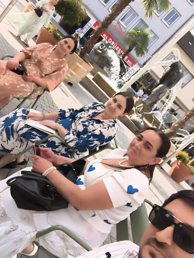 A group of people are photographed outdoors, possibly in a town square, with a fountain in the background. The people appear to be in casual or semi-formal attire. Two women are seated in chairs, one wearing a light-colored dress with a floral pattern, and the other wearing a blue and white patterned dress. Both women are looking towards the camera. A third person, a man, is in the foreground, also looking at the camera. In the background, a woman in a light green dress is walking away from the camera. The environment suggests a sunny day in a European city, possibly Heilbronn, Germany, given the architecture and fountain, which resemble those found in European squares.