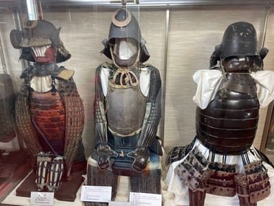 The image displays three suits of traditional Japanese samurai armor displayed in a museum setting. Each suit is mounted on a mannequin, showcasing the intricate craftsmanship and protective elements characteristic of samurai warfare. The armor on the left is a full suit, featuring a distinctive red and black lacquered breastplate and segmented leg guards. The central suit is also complete, with a dark, weathered helmet and a chainmail-like construction on the upper body. The third suit on the right is partially visible, with a dark helmet and a segmented chest plate over white fabric. The background appears to be a neutral, textured wall, possibly designed to highlight the artifacts. Small information cards are visible in front of the mannequins, likely providing details about the armor's origin and historical significance. One card in the foreground mentions "Iga City Education Committee," indicating the location as Iga, Japan.