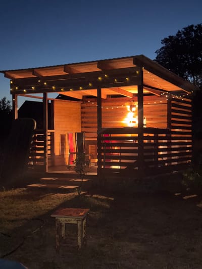 The image shows an outdoor wooden gazebo at dusk, illuminated by string lights and an interior light source. The gazebo has a slatted exterior and roof structure. Inside, there are two chairs with striped upholstery and a table. A bottle of water is visible on the table. In front of the gazebo, on the grass, there is a small, weathered stool and a coiled black hose. The background shows trees and a dark sky. The scene is set in the Ганчарскі сельскі Савет, Belarus. There are no people or animals visible in the image.