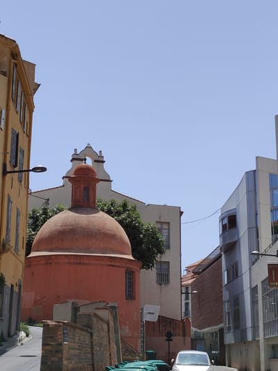 The image shows a view of the city of Perpignan, France, on a clear, sunny day. In the foreground, a prominent orange dome is visible, which is part of the Église Saint-Jacques. The dome is octagonal at its base and tapers to a smaller octagonal section topped with a finial. The church's facade, with its distinctive bell tower and arched openings, can be seen behind the dome.
To the left of the dome, a yellow building with dark shutters on its windows is partially visible. A black street lamp is mounted on this building.
To the right of the dome, several modern buildings are present. One building has a curved, metallic facade with multiple floors and large windows. Another building, constructed with red brick, juts out horizontally.
In the background, the sky is a clear, light blue. The street in the foreground is paved with asphalt and appears to be a gentle incline. There are several green trash bins to the left of the frame and a white car is parked on the street in the center-right. The overall impression is a view of a street in a city with a mix of historical and modern architecture.