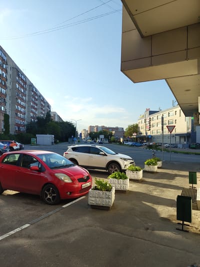 The image depicts a street scene in Zhukovsky, Russia, with a view of a long, multi-story apartment building on the left and a modern building on the right. In the foreground, a red hatchback is parked, and behind it, a white SUV is also parked. Several rectangular planters with greenery are placed along the sidewalk. The street in the background shows traffic with cars and a bus. The sky is clear blue with some thin power lines visible. The architecture of the buildings suggests a post-Soviet urban environment. There are no prominent actions or interactions visible, and the scene appears to be a typical daytime street view.