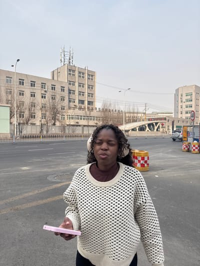 This image depicts an urban street scene in Dadong District, Shenyang, China, under a clear sky. A Black female, approximately adult, stands in the foreground, slightly right of center, with her eyes closed or squinted and lips pursed. She wears a cream-colored, textured knit sweater over a dark brown top, and light-colored headphones or earmuffs cover her ears. In her left hand, she holds a light pink rectangular object, consistent with a smartphone. The background features a multi-lane asphalt road. To the left, a grey multi-story building with numerous windows is visible; Chinese characters on its facade identify it as the "Shenyang University Student Innovation and Entrepreneurship Center." Multiple communication antennae are mounted on its roof. In front of this building are bare trees and a metal fence. To the right, a series of orange and white checkered traffic barriers are positioned along the roadside. Further in the background, a distinctive bridge with red structural elements spans across a thoroughfare, and another grey building is visible. A white SUV is present in the distance near the bridge, and a circular "40" speed limit sign is visible on the right side of the road.