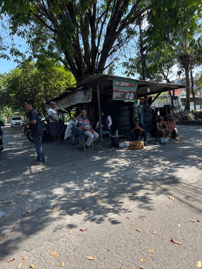 This media file depicts an outdoor scene in Sepande, Indonesia, likely a tire repair shop or a similar roadside establishment. The image is oriented vertically. Several men are present, some seated and appearing to rest or wait. One man is standing and appears to be leaning against a motorcycle. Another man is seated inside the shaded structure, surrounded by stacks of tires. A sign is visible on the left side of the structure, with the text "KSN BAN" prominently displayed in green and white. Beneath this, there appears to be more text, possibly advertising or branding.

The setting is a street, with asphalt paving and scattered dry leaves. The scene is illuminated by bright sunlight, casting distinct shadows from trees and the overhead shelter, suggesting it is daytime, likely mid-morning or afternoon. Lush green trees and foliage are visible, particularly to the left and above the structure, indicating a tropical or sub-tropical environment. The overall atmosphere seems relaxed, with the individuals appearing to be taking a break from work. No specific activity other than resting or waiting is clearly evident. The presence of tires, a compressor, and a motorcycle suggests an automotive-related service.