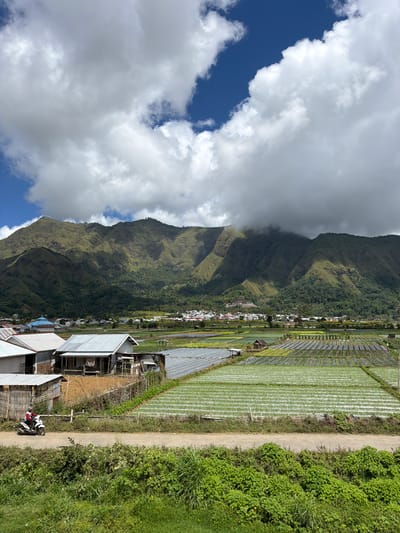 The image shows a mountainous landscape in Sembalun Bumbung, Indonesia. The left side features a large mountain partially obscured by clouds, revealing blue sky. A line of buildings and vegetation extends from the mountain base to the lower right. Adjacent to this line are extensive rice paddies. A dirt path runs along the right side, bordered by dense green vegetation. A building and a motorcycle are visible in the foreground along the path.