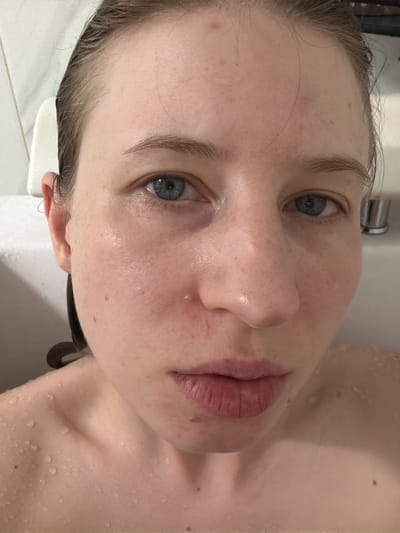A close-up, waist-up, head-on shot shows a young woman with wet, light brown hair slicked back, revealing her forehead and face. She is looking directly at the camera with her blue eyes. Her skin is pale with visible freckles and appears slightly shiny, likely from water. There are small blemishes on her forehead and cheek. Her lips are slightly parted, revealing a few light-colored bumps. Water droplets are visible on her shoulders and décolletage. The background is out of focus, but appears to be a tiled bathroom environment, with a white bathtub edge visible on the left and a chrome fixture on the right. The lighting is soft and appears to come from above. The overall impression is one of a candid, personal moment, possibly taken in a bathroom or shower. This image was captured in Dongducheon-si, South Korea.